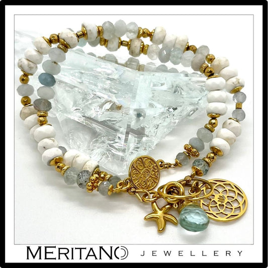 Biała, podwójna bransoletka z howlitem i akwamarynem, ozdobiona złotą monetą obok szkła Biała, podwójna bransoletka z howlitem i akwamarynem, ozdobiona złotą monetą obok szkła