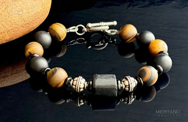 Bransoletka męska z kamieni turmalinu tygrysie oko, leżąca na czarnym tle w towarzystwie pomarańczowego kamienia