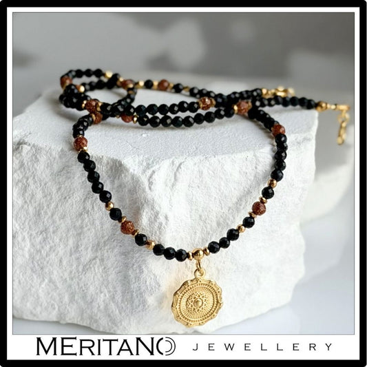 Naszyjnik z czarnego turmalinu i piasku pustyni z okrągłą złotą zawieszką ułożony na jasnym, surowym kamieniu ekspozycyjnym